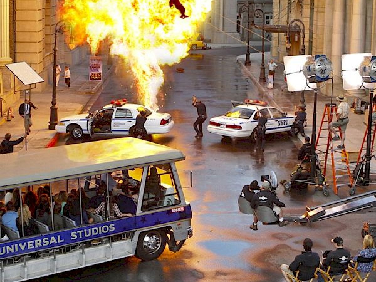 This image shows a film set with an explosion, police cars, cameras, crew, and a Universal Studios tour tram loaded with people under a bright light.