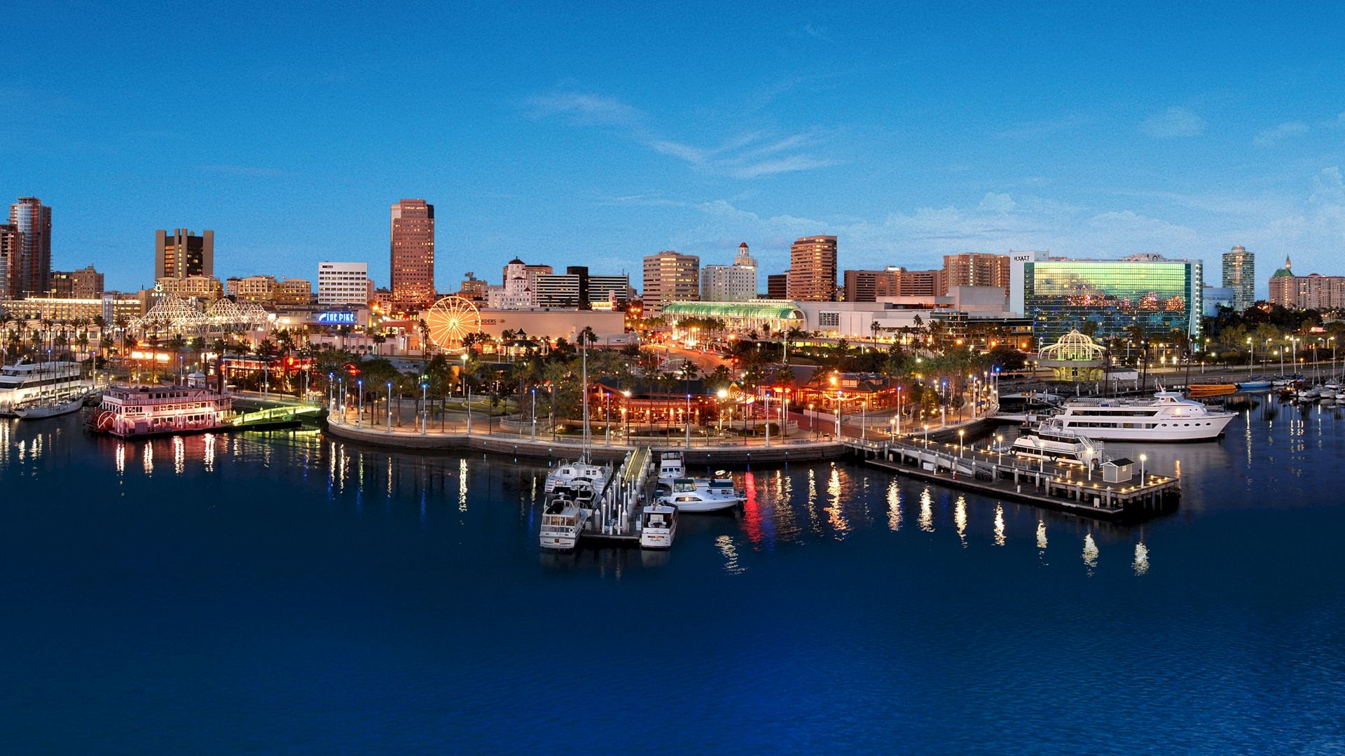 The image shows a vibrant waterfront cityscape at twilight, featuring illuminated buildings, a marina with docked boats, and a calm body of water.