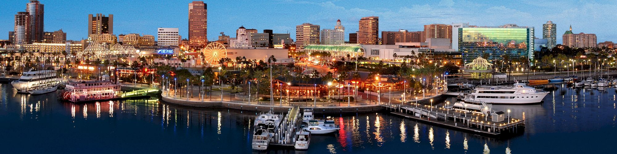 The image shows a vibrant waterfront cityscape at twilight, featuring illuminated buildings, a marina with docked boats, and a calm body of water.