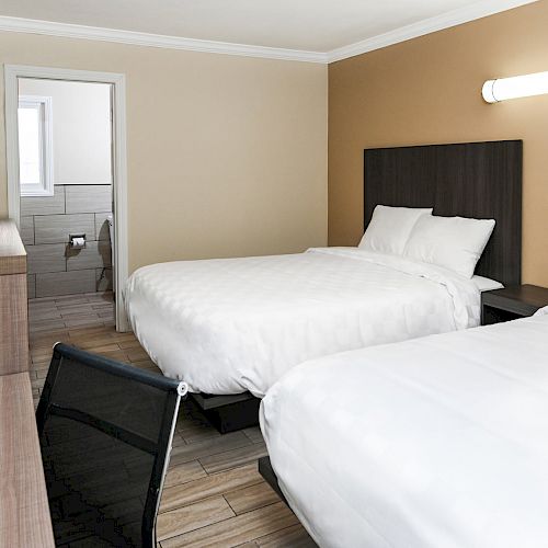 A hotel room with two double beds, a TV, a desk, a chair, and a bathroom with a partially opened door, displaying modern, minimalistic decor.