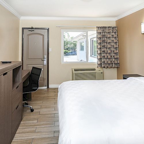 A modern hotel room featuring a large bed, a wall-mounted TV, a desk with a chair, and a window with patterned curtains.