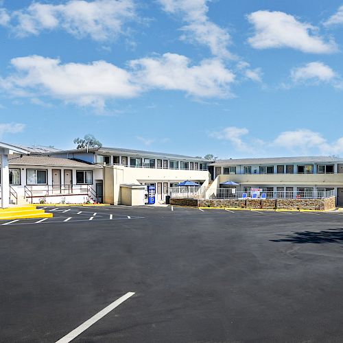 The image shows a modern motel with multiple buildings, a parking lot with marked spaces, and a clear blue sky above.