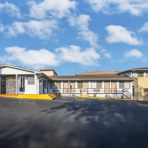 The image shows the exterior of a building complex under a blue sky with scattered clouds, featuring parking space and a blue vending machine on the right.