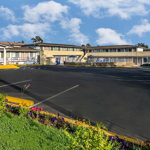 The image shows a building complex with a large, empty parking lot in front, surrounded by greenery and flowering plants under a bright blue sky.