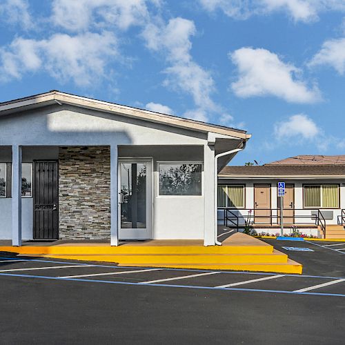 The image shows a single-story building with a covered entrance, ramp, and adjacent parking spaces under a partly cloudy sky.