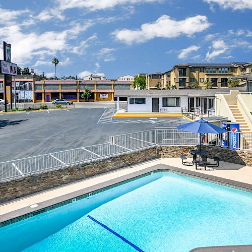 Outdoor scene of a motel-style lodging with a parking lot, small swimming pool, lounge chairs, umbrellas, and a few multi-story buildings around.