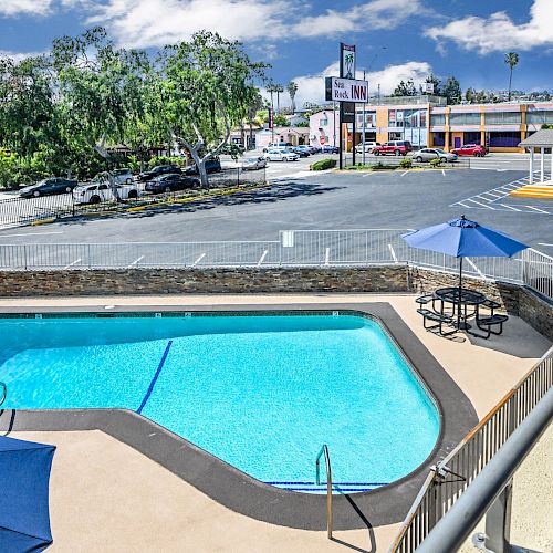 The image shows an outdoor pool area with umbrellas and chairs, surrounded by a fence. There is a parking lot and streets visible in the background.