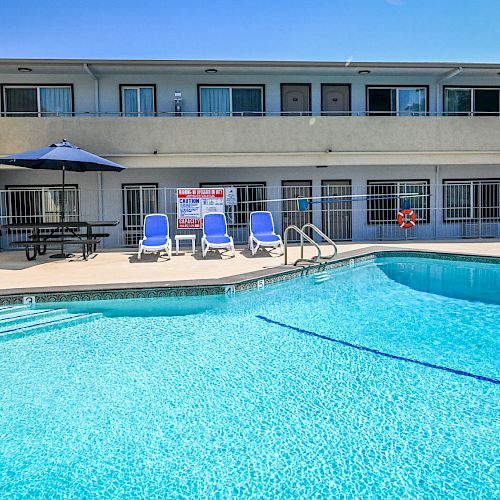 The image shows a swimming pool in front of a two-story building, with blue lounge chairs and umbrella. There is a sign on the building's railing.