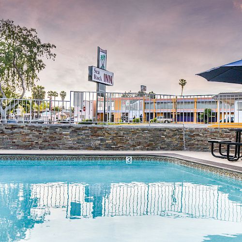 An outdoor swimming pool with clear blue water, surrounded by a stone wall. There's also a picnic table with an umbrella and a sign in the background.