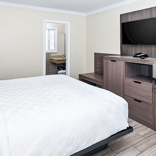 A hotel room with a large bed, TV, desk, chair, and visible bathroom. Light decor with wooden furnishings and minimalistic design.