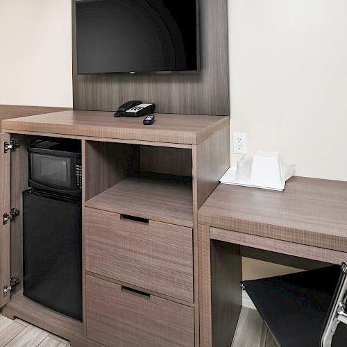 The image shows a desk with a mounted TV above it, featuring a small fridge, microwave, telephone, drawers, and a chair at the desk.