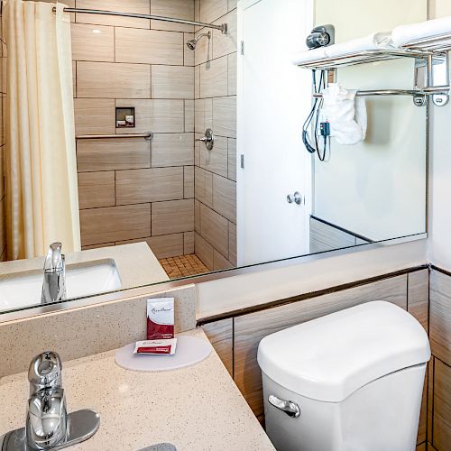 A modern bathroom with a sink, mirror, toilet, and a shower area enclosed by a curtain. Towels and toiletries are neatly arranged.