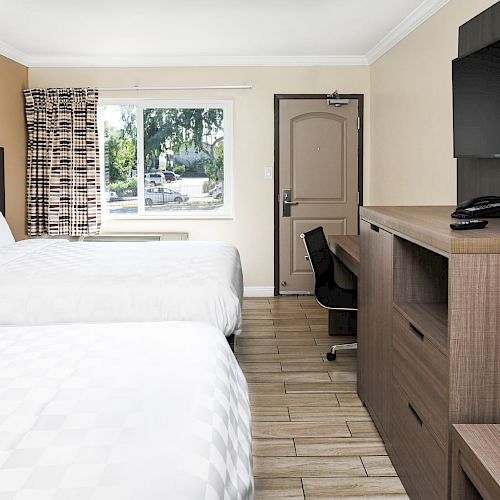 A modern hotel room with two beds, a TV, desk, and dresser, featuring a window with a view and a door.