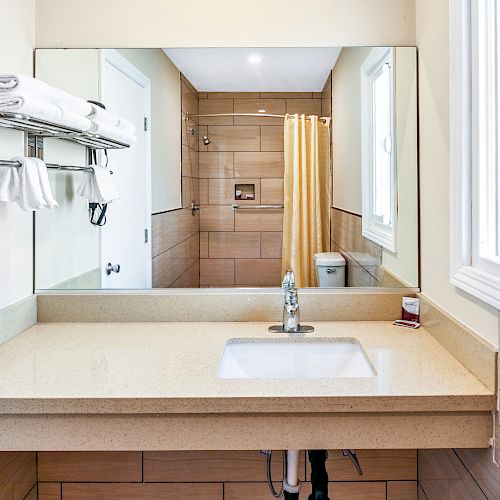 A bathroom with a countertop sink, mirror, wall-mounted hairdryer, towel shelf, and shower area with a yellow curtain is shown.