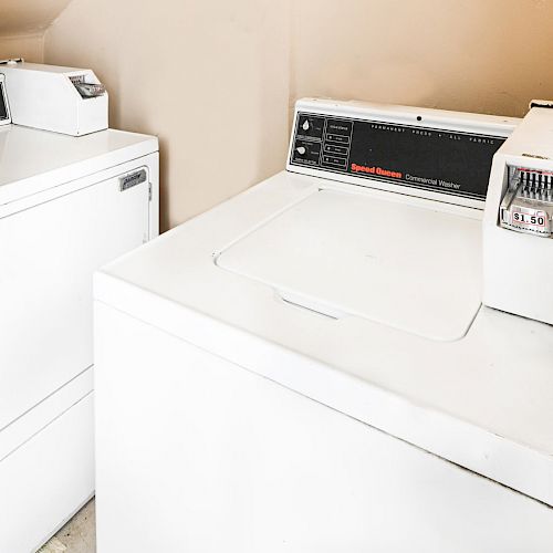 The image shows a laundry room with a white washing machine and a white dryer, each equipped with coin-operated controls.