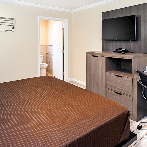 This image shows a tidy hotel room with a brown bedspread, TV on a wall-mounted cabinet, desk and chair, and a view into a bathroom.