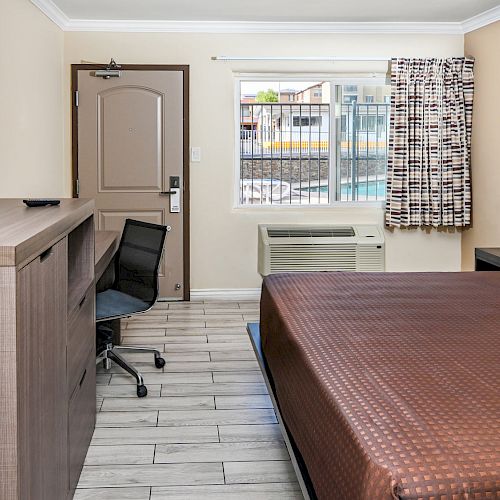 The image shows a tidy hotel room with a bed, desk, chair, TV, and a small window. There is also an air conditioning unit beneath the window.