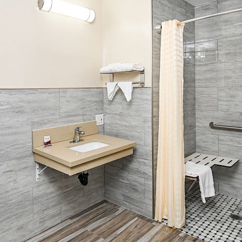 A modern bathroom with a walk-in shower, foldable shower seat, sink, grab bar, towels, and a window. The bathroom has grey tiles and wood flooring.