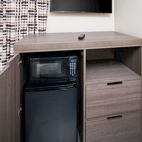 The image shows a cabinet with a mini refrigerator and two drawers. A remote rests on top of the cabinet, with a mounted TV and patterned curtains nearby.