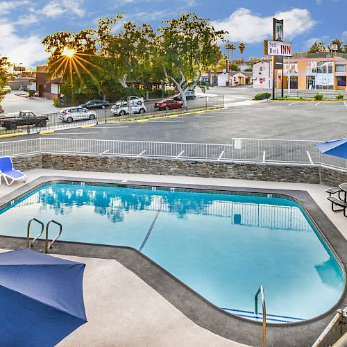 The image shows a pool area with blue umbrellas and lounge chairs, adjacent to a parking lot and nearby buildings under a partly cloudy sky.