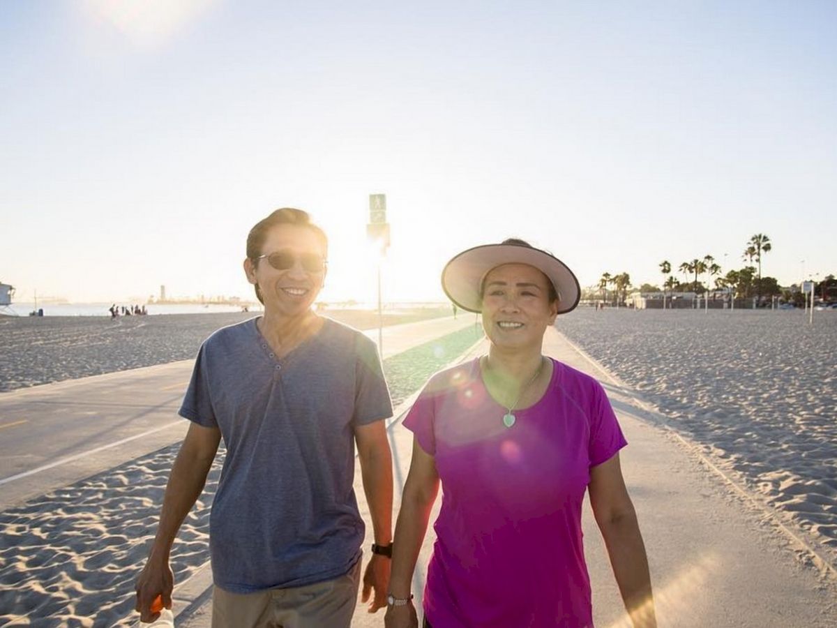 A man and woman are walking on a beach path, smiling, with the sun setting in the background. The beach is sandy with sparse palm trees.