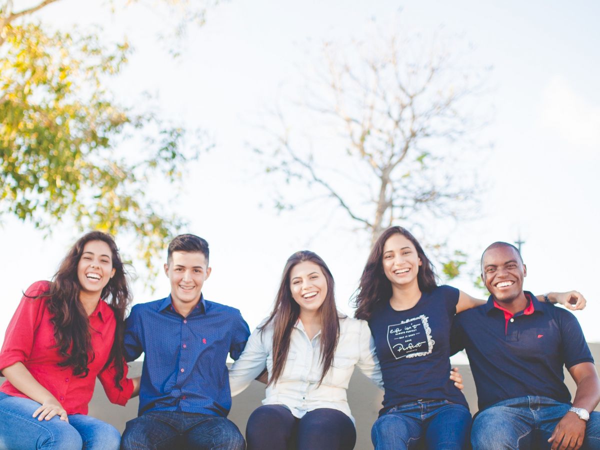 A group of five people sitting on a ledge outdoors, smiling and enjoying each other's company, with trees and a clear sky in the background.