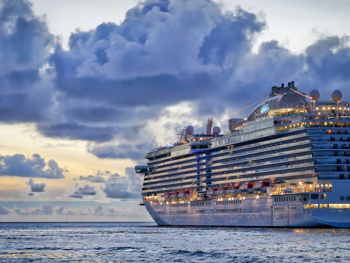 A large cruise ship sails on the ocean at dusk, with clouds overhead and calm waters below, creating a serene and picturesque scene.