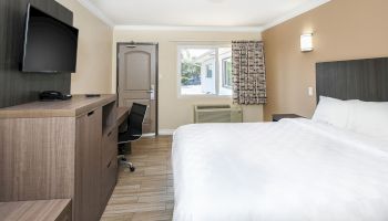 The image shows a modern hotel room with a bed, a wall-mounted TV, a desk chair, wooden furniture, a window with curtains, and a door.