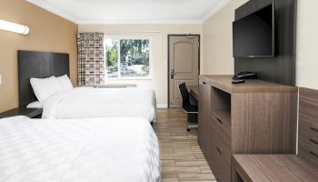 The image shows a hotel room with two beds, a TV on a dresser, a desk with a chair, and a window near a door. The room is simply furnished.