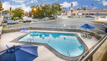 The image shows a small outdoor swimming pool with two blue umbrellas, lounge chairs, and a picnic table in a fenced area near a parking lot.