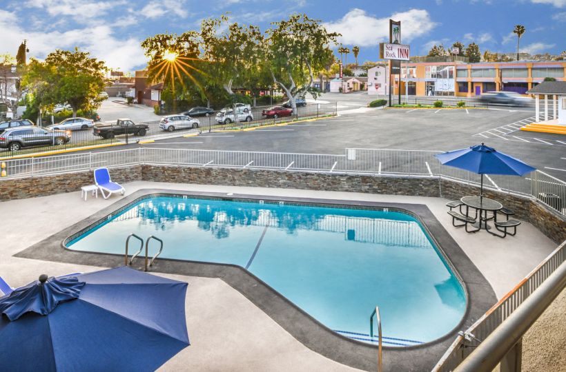 The image shows a small outdoor swimming pool with two blue umbrellas, lounge chairs, and a picnic table in a fenced area near a parking lot.