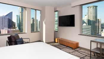 A modern bedroom with large windows showcasing a city view, a wall-mounted TV, and minimalistic furniture including a chair and a small table.