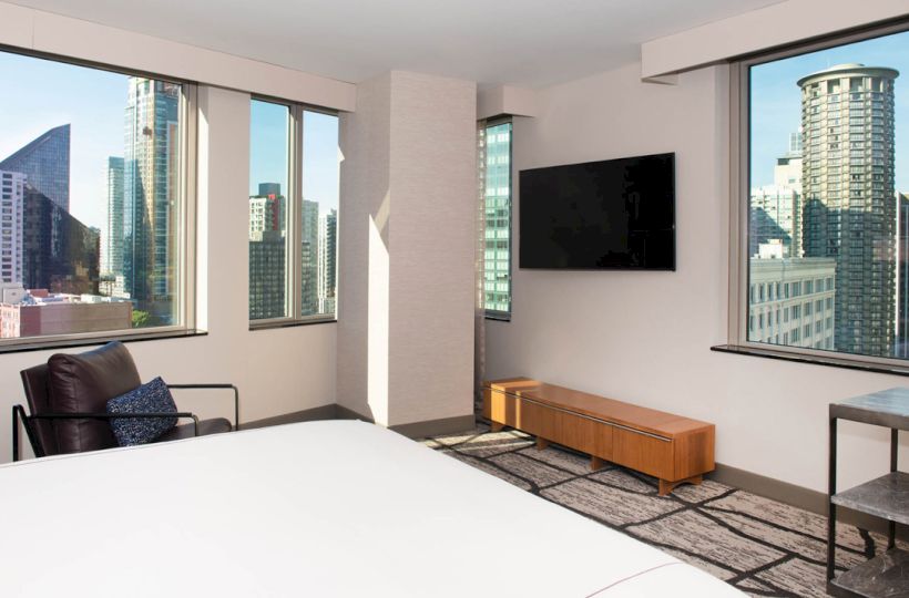 A modern bedroom with large windows showcasing a city view, a wall-mounted TV, and minimalistic furniture including a chair and a small table.