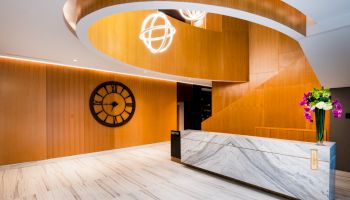 A modern interior area with a marble reception desk, wooden panels, a large clock, stylish lighting, and a vase of colorful flowers.