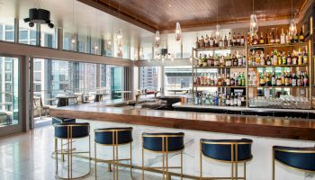 The image shows a modern bar with high shelves stocked with various bottles of alcohol, blue bar stools, and large windows offering a city view.