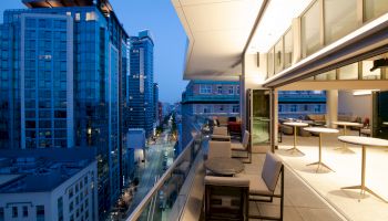 A modern cityscape at dusk viewed from a balcony with seating and tables, adjacent to a brightly lit interior space.