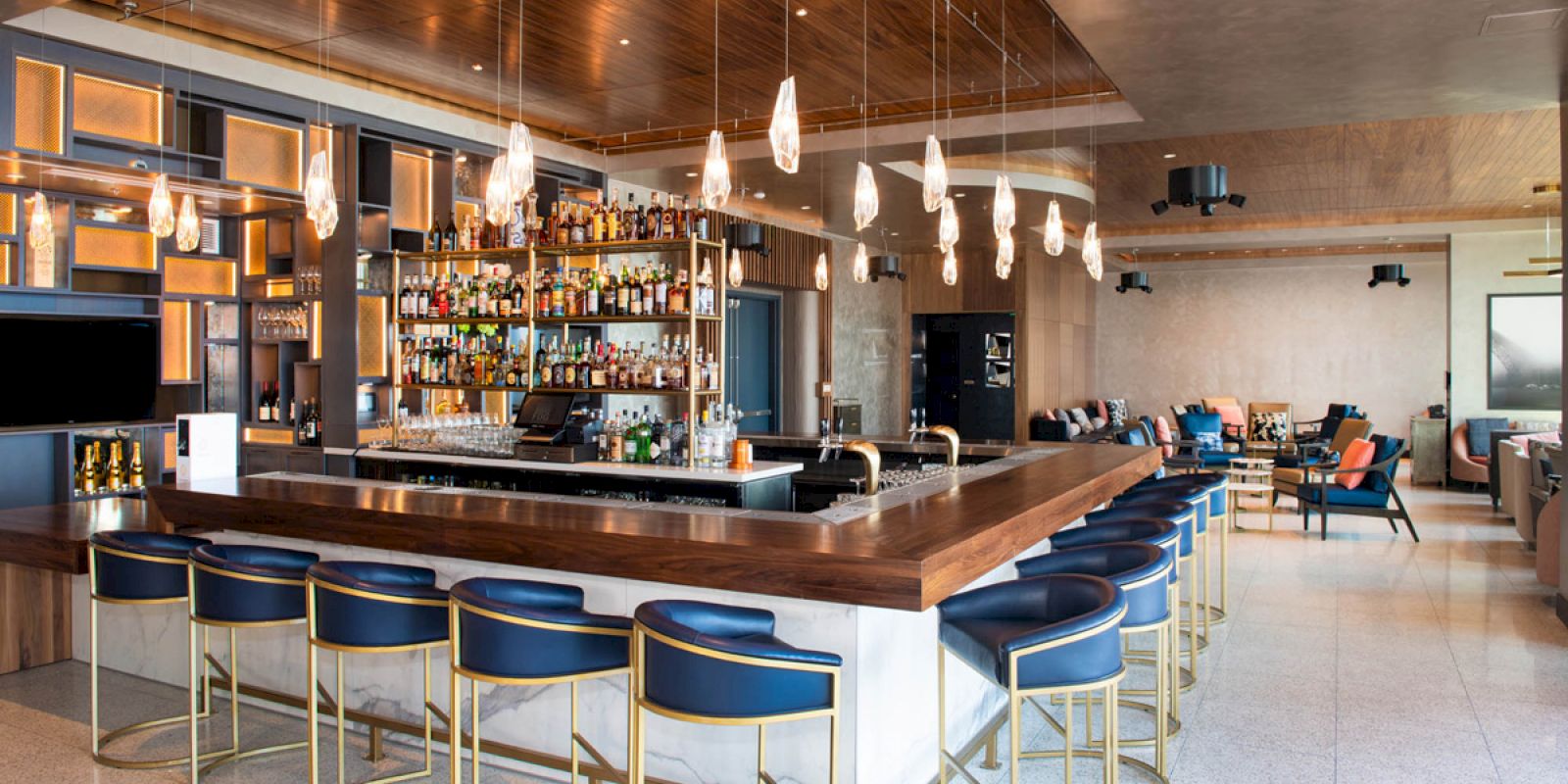 The image shows a modern bar with blue stools, a wooden counter, hanging pendant lights, and a well-stocked bar area. The seating area has a cozy ambiance.