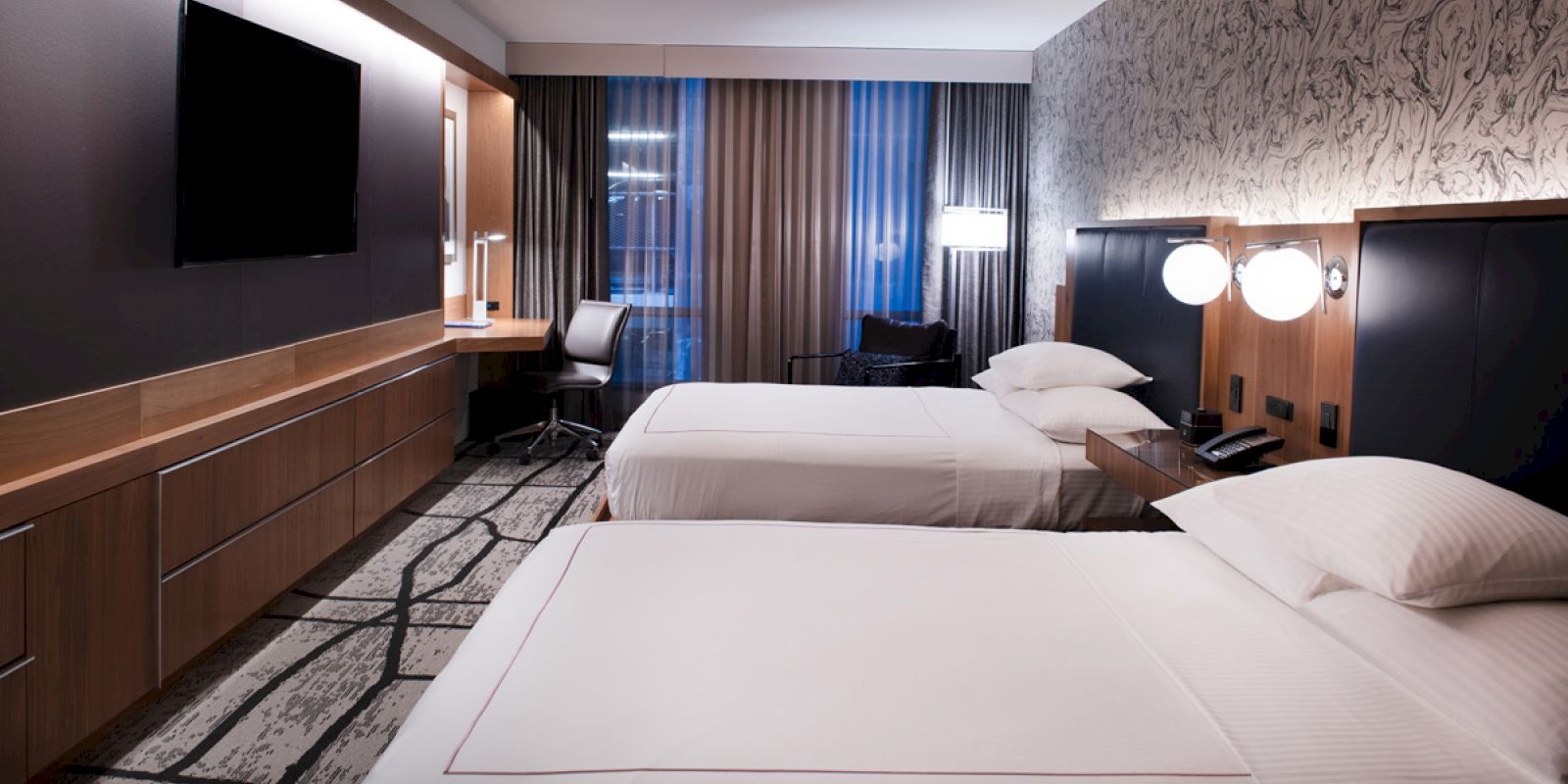 The image shows a modern, well-lit hotel room with two double beds, a desk, a wall-mounted TV, and a window with curtains ending the sentence.