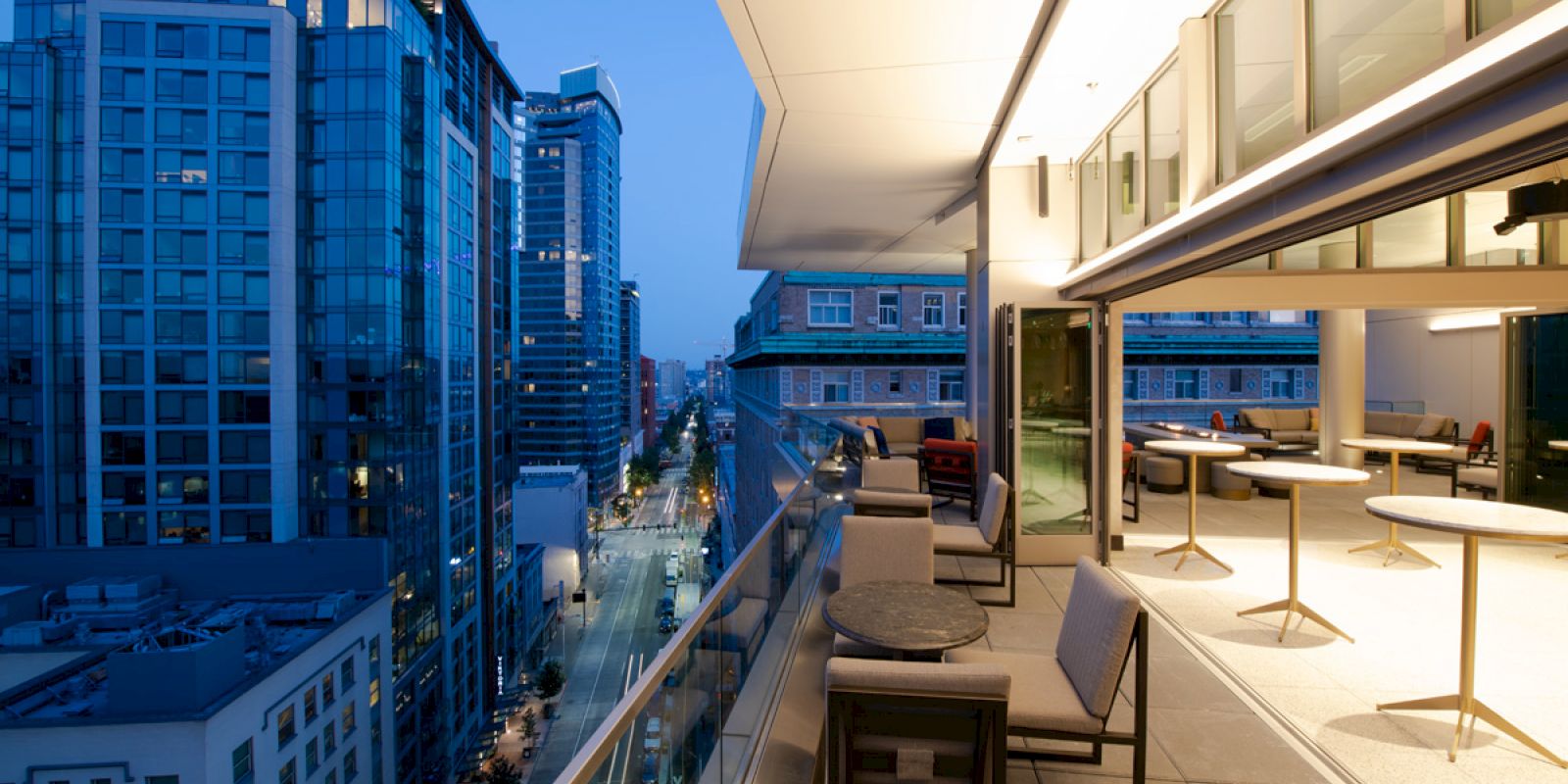 A modern cityscape with a view from a stylish rooftop patio, featuring seating and tables, overlooking buildings and a street at dusk.