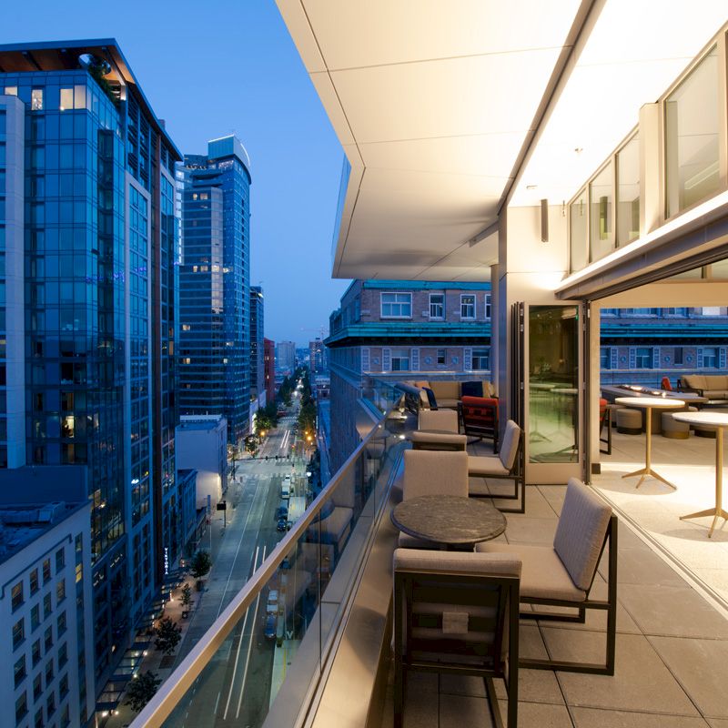 A modern urban balcony with seating overlooks a cityscape with tall buildings and a street below, illuminated by evening lights.