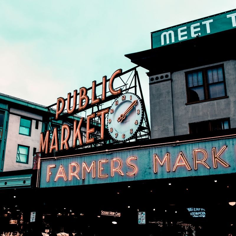 The image displays a vibrant neon sign for a Public Market and Farmers Market with a clock, set against buildings and a visible "Meet the" sign.