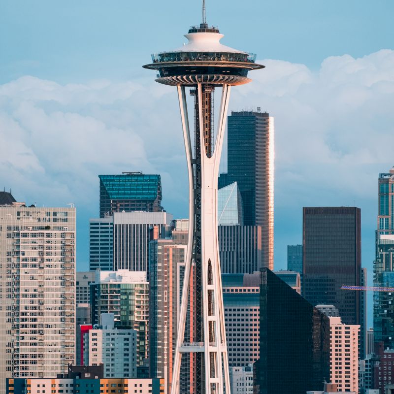 The image features a skyline with the Space Needle prominently, surrounded by various modern buildings under a blue sky with light clouds, creating an urban scene.