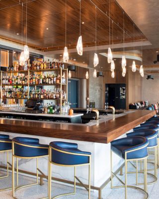 The image shows a modern bar with a wooden counter, blue barstools, hanging lights, and a fully stocked shelf of liquor bottles behind the bar.