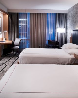 The image shows a modern hotel room with two neatly made beds, a wall-mounted TV, a desk, and a chair next to a window with curtains.