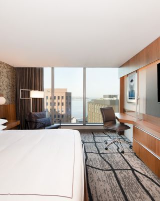 This image shows a modern hotel room with a large bed, a desk and chair, a flat-screen TV, and a window offering a cityscape view.