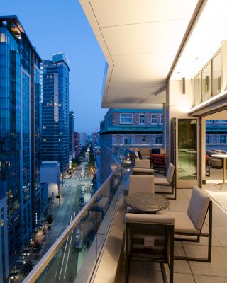 A modern cityscape at dusk with high-rise buildings, viewed from a spacious balcony featuring outdoor seating and tables.