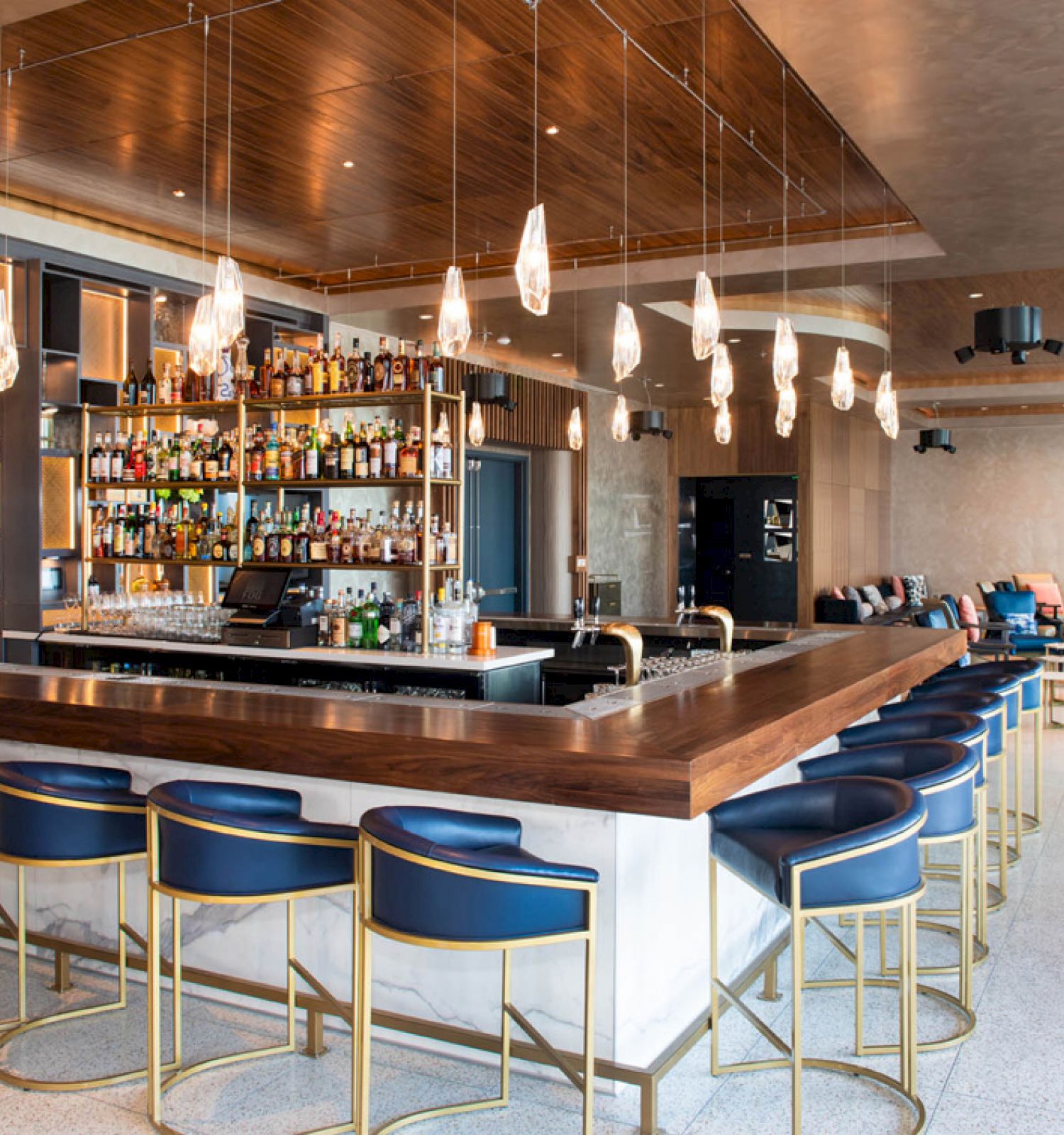 The image shows a modern bar with blue bar stools, a well-stocked liquor shelf, hanging lights, and a cozy lounge area with sofas and chairs.