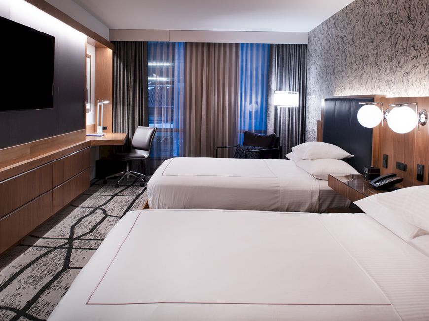 A modern hotel room with two double beds, a flat-screen TV, a desk with a chair, and curtains covering the windows.