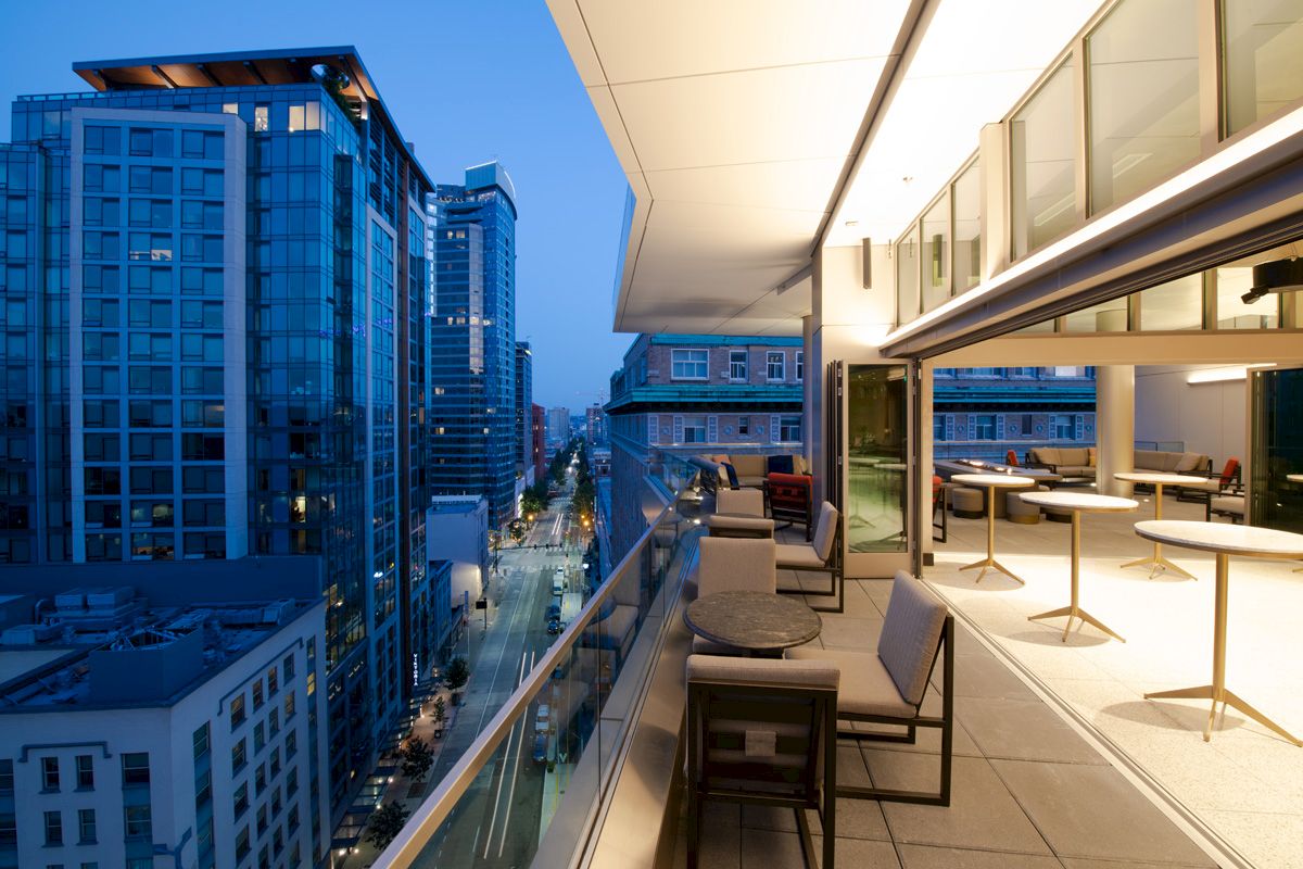 A modern outdoor balcony at twilight, with table and chairs, overlooks a city street lined with tall, illuminated buildings.