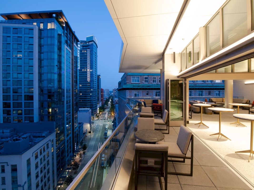 A modern outdoor balcony at twilight, with table and chairs, overlooks a city street lined with tall, illuminated buildings.